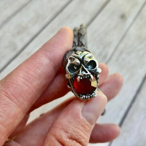 May include: A silver skull bracelet with a red gemstone in the mouth. The bracelet is made of braided leather.