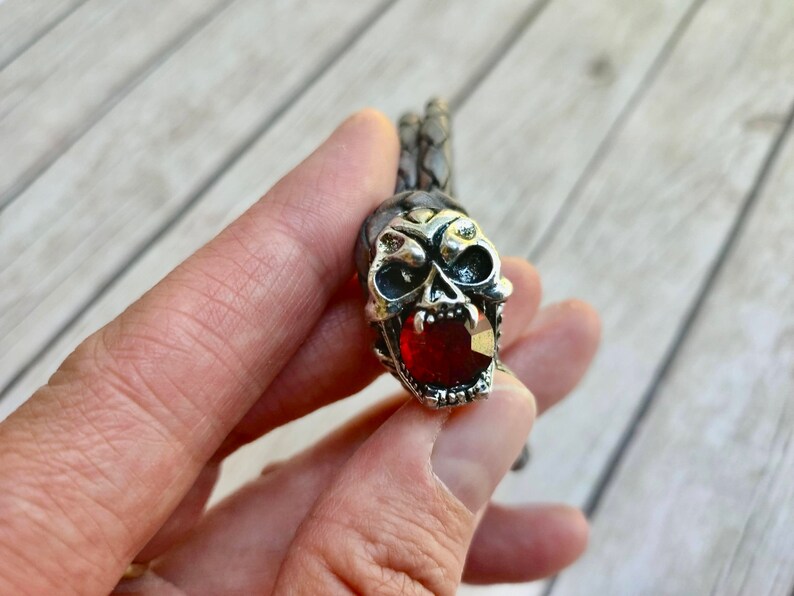 May include: A silver skull bracelet with a red gemstone in the mouth. The bracelet is made of braided leather.