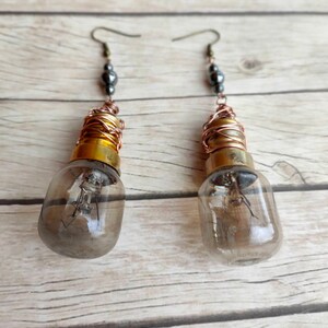 May include: A pair of dangle earrings featuring small light bulb pendants wrapped in copper wire. Each bulb has a gold-colored base and a clear glass casing. The earrings are accented with black beads.