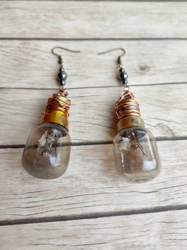 May include: A pair of dangle earrings featuring small light bulb pendants wrapped in copper wire. Each bulb has a gold-colored base and a clear glass casing. The earrings are accented with black beads.