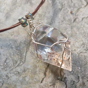May include: A clear, faceted crystal pendant wrapped in silver-toned wire, suspended from a brown cord. The pendant features two small, iridescent, cylindrical beads at the top where the cord meets the wire-wrapped crystal point.
