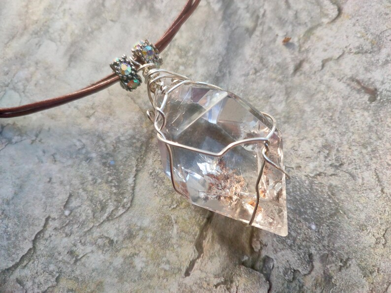 May include: A clear, faceted crystal pendant wrapped in silver-toned wire, suspended from a brown cord. The pendant features two small, iridescent, cylindrical beads at the top where the cord meets the wire-wrapped crystal point.