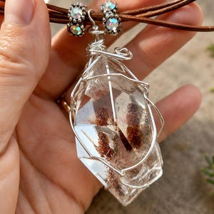 May include: A clear, faceted quartz crystal pendant with internal reddish-brown inclusions, encased in a silver-toned wire wrap. The pendant hangs from a brown leather cord adorned with two silver-toned beads featuring small, iridescent crystals.
