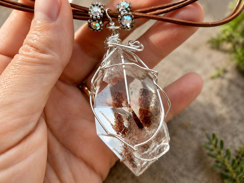 May include: A clear, faceted quartz crystal pendant with internal reddish-brown inclusions, encased in a silver-toned wire wrap. The pendant hangs from a brown leather cord adorned with two silver-toned beads featuring small, iridescent crystals.
