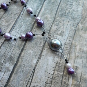 Peut inclure: Un collier en argent avec un grand pendentif en perle grise et de plus petites perles violettes et blanches. Le collier est sur une surface en bois.