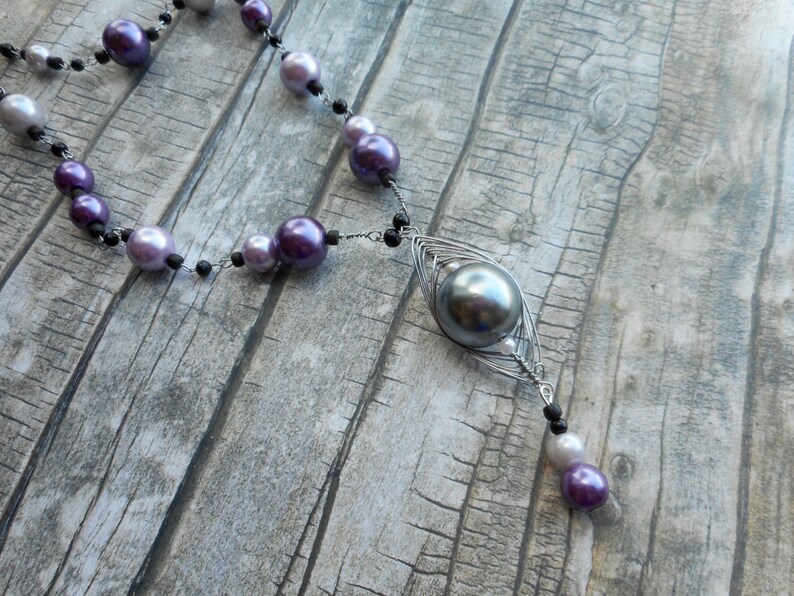 Peut inclure: Un collier en argent avec un grand pendentif en perle grise et de plus petites perles violettes et blanches. Le collier est sur une surface en bois.