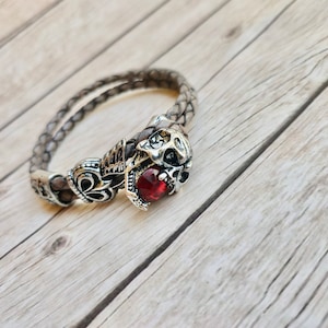May include: A brown leather bracelet featuring a silver skull charm with a red gemstone. The bracelet is braided and has an intricate design.