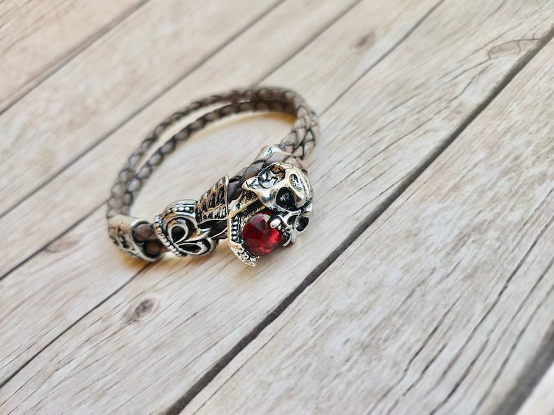 May include: A brown leather bracelet featuring a silver skull charm with a red gemstone. The bracelet is braided and has an intricate design.
