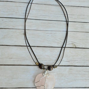 May include: A pink heart-shaped pendant necklace with a silver wire design. The necklace has a brown leather cord with brown wooden beads.