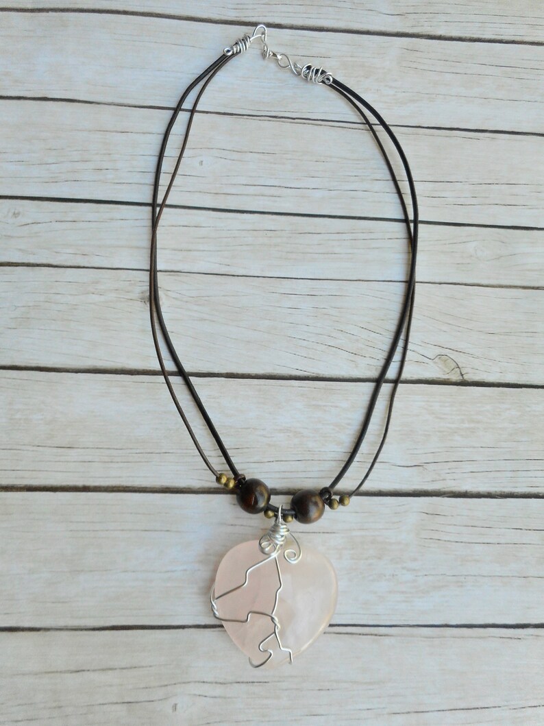 May include: A pink heart-shaped pendant necklace with a silver wire design. The necklace has a brown leather cord with brown wooden beads.