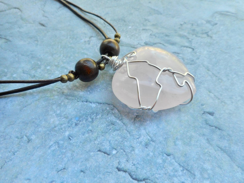 May include: A pink rose quartz heart pendant wrapped in silver wire, hanging from a brown leather cord with wooden beads.