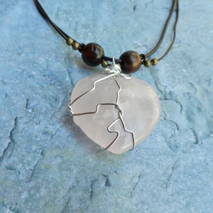 May include: A pink heart-shaped rose quartz pendant wrapped in silver wire, hanging from a brown leather cord with wooden beads.