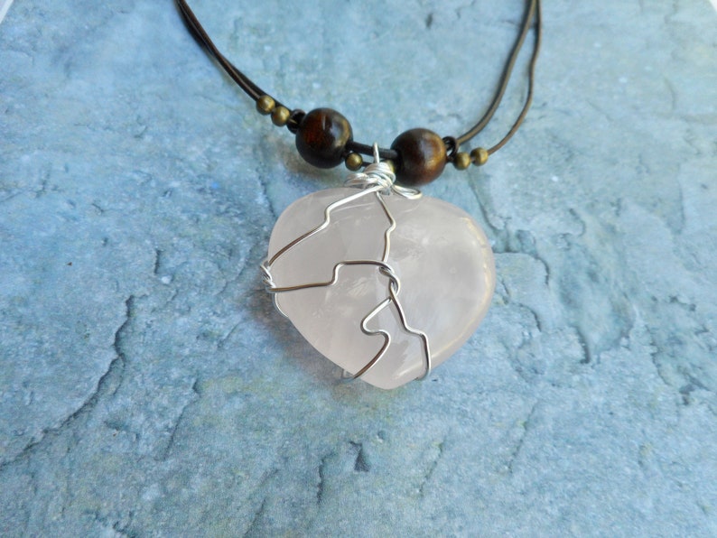 May include: A pink heart-shaped rose quartz pendant wrapped in silver wire, hanging from a brown leather cord with wooden beads.