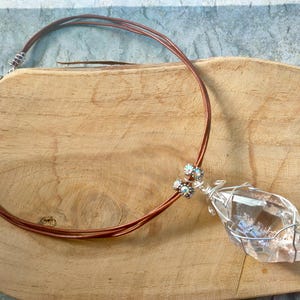 May include: A necklace featuring a clear, faceted crystal pendant wrapped in silver-toned wire. The pendant is attached to a multi-strand brown leather cord, accented with small, sparkling, multi-coloured decorative beads near the crystal attachment.