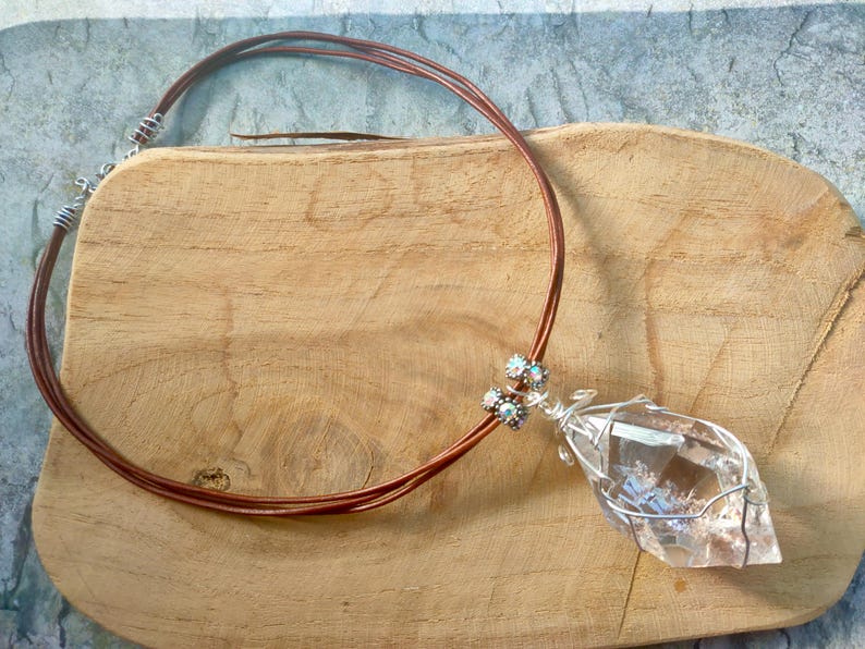 May include: A necklace featuring a clear, faceted crystal pendant wrapped in silver-toned wire. The pendant is attached to a multi-strand brown leather cord, accented with small, sparkling, multi-coloured decorative beads near the crystal attachment.