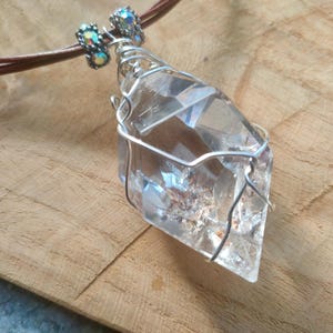 May include: A clear, faceted crystal pendant wrapped in silver-toned wire, suspended from a brown leather cord. The necklace features two small, iridescent, cylindrical beads with metallic accents positioned above the crystal point.