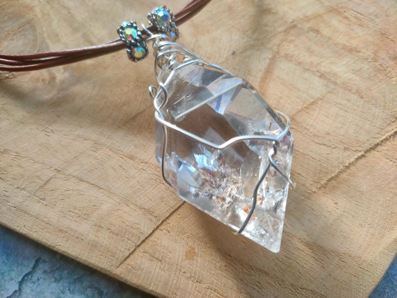 May include: A clear, faceted crystal pendant wrapped in silver-toned wire, suspended from a brown leather cord. The necklace features two small, iridescent, cylindrical beads with metallic accents positioned above the crystal point.