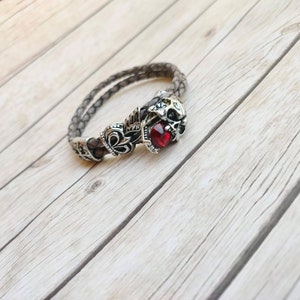May include: A grey leather bracelet with a silver skull charm and a red gemstone. The bracelet is braided and features a decorative design.