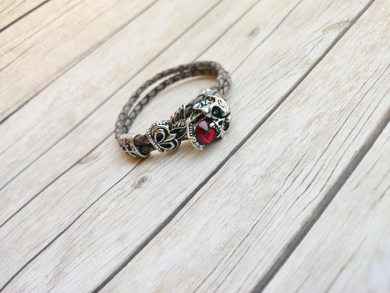 May include: A grey leather bracelet with a silver skull charm and a red gemstone. The bracelet is braided and features a decorative design.