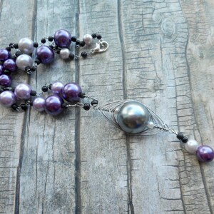Peut inclure: Un collier multi-rangs avec des perles violettes, blanches et noires. Le collier a un grand pendentif en perle grise avec un design de cage en fil m&eacute;tallique.