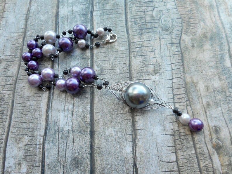 Peut inclure: Un collier multi-rangs avec des perles violettes, blanches et noires. Le collier a un grand pendentif en perle grise avec un design de cage en fil m&eacute;tallique.