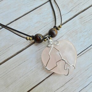 May include: A pink heart-shaped rose quartz pendant necklace with silver wire wrapping. The necklace has a brown leather cord with brown wooden beads.