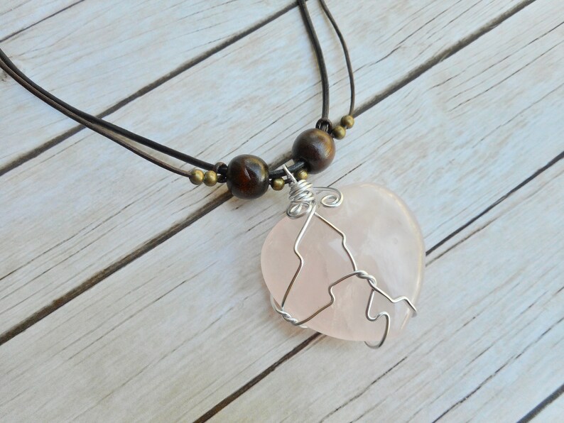 May include: A pink heart-shaped rose quartz pendant necklace with silver wire wrapping. The necklace has a brown leather cord with brown wooden beads.