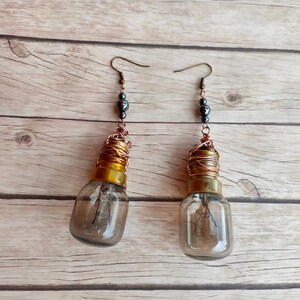 May include: A pair of earrings made from repurposed light bulbs. The bulbs are wrapped in copper wire and have a gold-colored top. The earrings are accented with black beads.