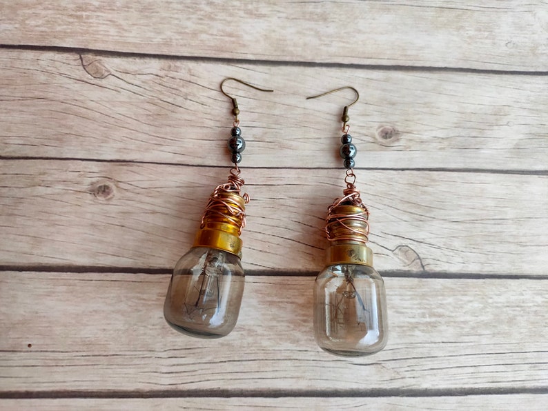 May include: A pair of earrings made from repurposed light bulbs. The bulbs are wrapped in copper wire and have a gold-colored top. The earrings are accented with black beads.