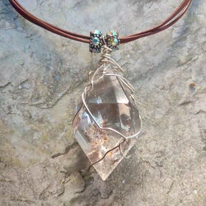 May include: A clear, faceted crystal pendant wrapped in silver-toned wire, suspended from a brown cord. The pendant features two small, metallic, iridescent beads at the top where the wire connects to the cord, showcasing a pointed, geometric shape.