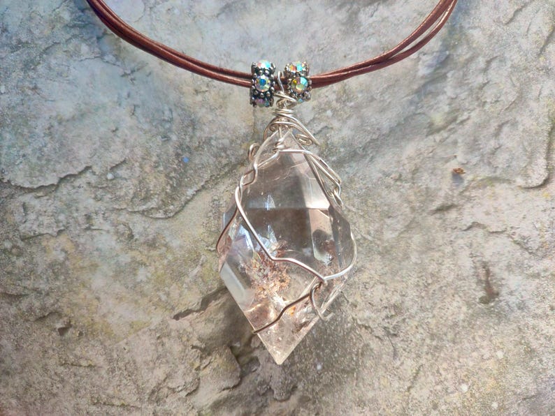 May include: A clear, faceted crystal pendant wrapped in silver-toned wire, suspended from a brown cord. The pendant features two small, metallic, iridescent beads at the top where the wire connects to the cord, showcasing a pointed, geometric shape.
