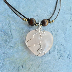 May include: A pink heart-shaped crystal pendant necklace with a silver wire wrap, hanging from a brown leather cord with wooden beads.