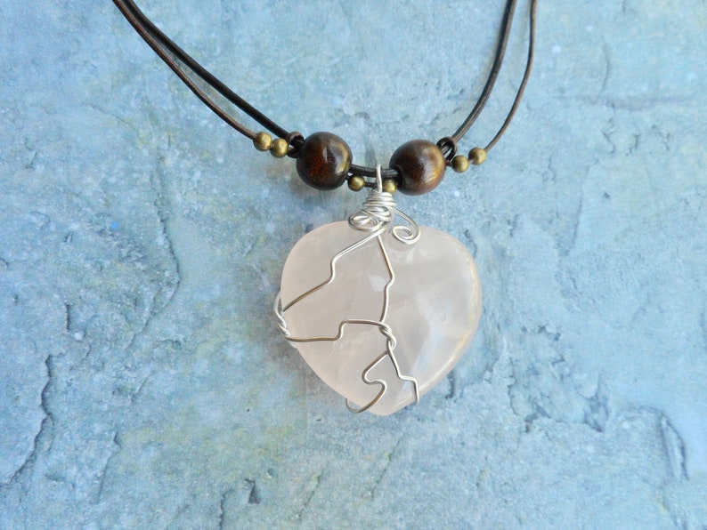May include: A pink heart-shaped crystal pendant necklace with a silver wire wrap, hanging from a brown leather cord with wooden beads.