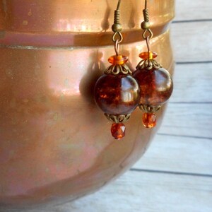 May include: A pair of amber glass earrings with gold-toned metal accents. The earrings feature a round, faceted amber glass bead with a smaller, clear glass bead dangling below.