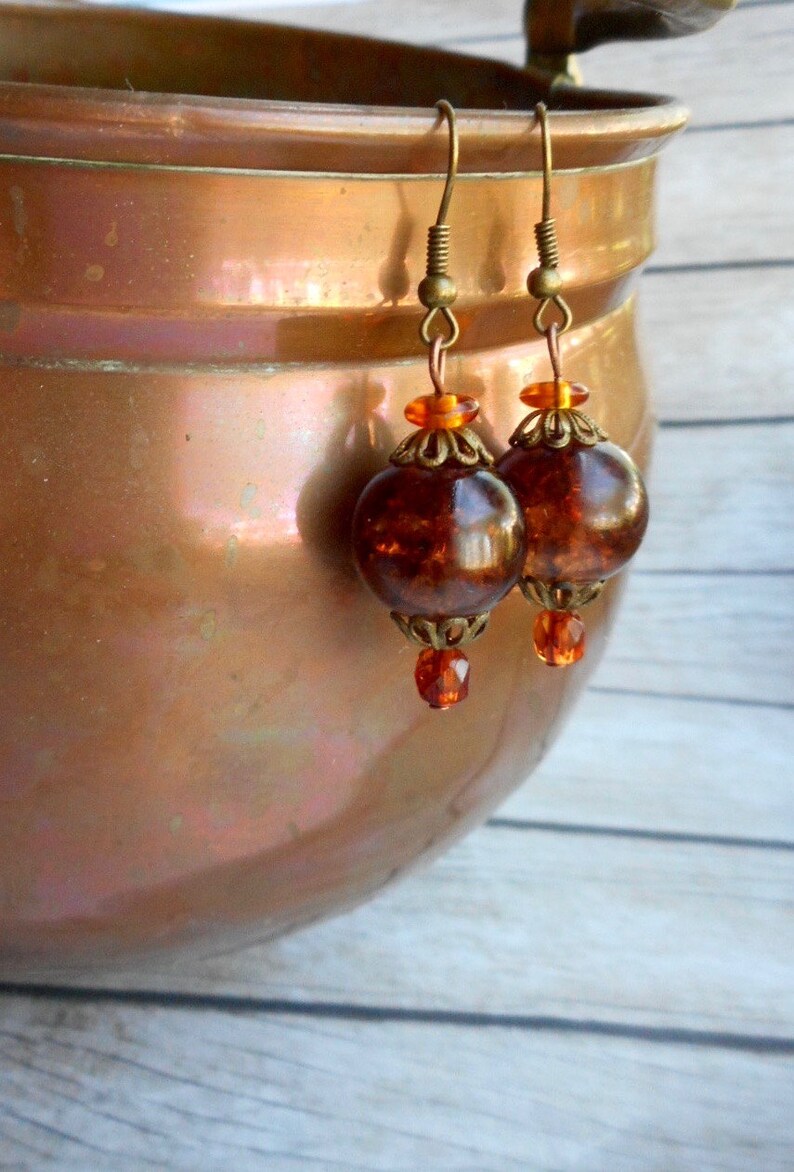 May include: A pair of amber glass earrings with gold-toned metal accents. The earrings feature a round, faceted amber glass bead with a smaller, clear glass bead dangling below.
