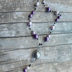 Peut inclure: Un collier en cha&icirc;ne argent&eacute;e avec un grand pendentif en perle gris fonc&eacute;. Le collier est compos&eacute; de perles violettes et blanches altern&eacute;es.
