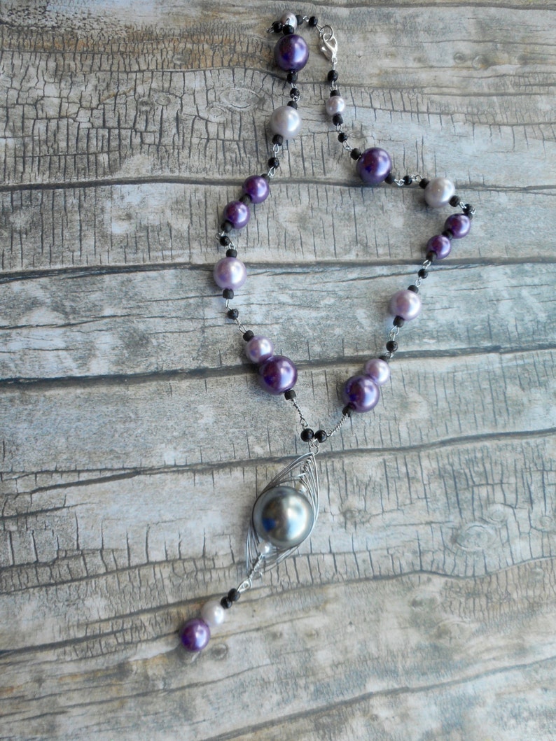 Peut inclure: Un collier en cha&icirc;ne argent&eacute;e avec un grand pendentif en perle gris fonc&eacute;. Le collier est compos&eacute; de perles violettes et blanches altern&eacute;es.
