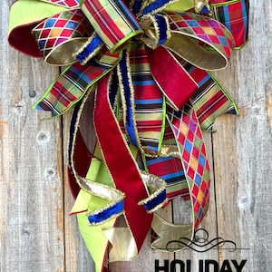 May include: A decorative bow featuring a mix of ribbons in solid red velvet, lime green, blue velvet, and multi-colored plaid and diamond patterns with gold metallic edges. The text HOLIDAY BAUBLES is printed in black on the lower right side.