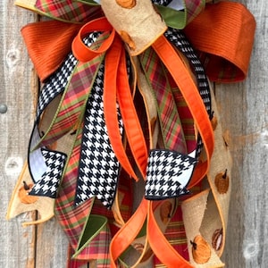 May include: A decorative bow featuring a mix of ribbons including tan burlap with embroidered orange pumpkins, black and white houndstooth, solid orange, and red, green, and gold plaid. The text HOLIDAY BAUBLES is printed at the bottom.