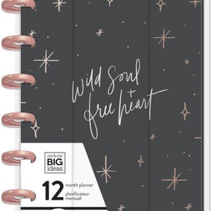 Peut inclure: Un agenda Happy Planner noir et rose gold avec un motif de nuit étoilée. L'agenda est un format horizontal de 12 mois et comporte le texte "Wild Soul + free heart".
