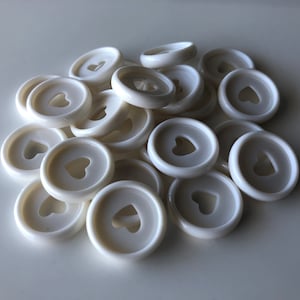 May include: A pile of white plastic heart-shaped buttons. The buttons are approximately 2.5 cm in diameter and have a small hole in the centre.