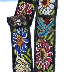 May include: Black beaded belt with colorful floral and bird designs. The belt features a repeating pattern of flowers in white, red, blue, and yellow, with green leaves. The intricate beadwork creates a detailed and vibrant accessory.