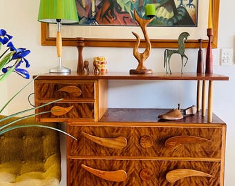 May include: A mid-century modern wooden dresser with three drawers and unique teardrop-shaped pulls. The dresser is adorned with decorative items, including a green lampshade, artwork, and a small bronze horse statue. A small rug is placed in front of the dresser.