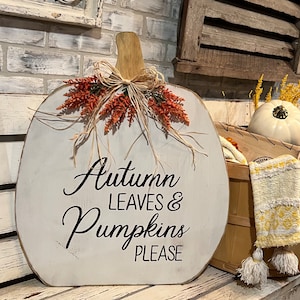 Könnte beinhalten: Ein weißes, hölzernes Kürbis-Schild mit dem Text "Autumn Leaves & Pumpkins Please" in schwarzen Buchstaben. Der Kürbis ist mit einer braunen Schleife und orange-braunem Herbstlaub verziert.