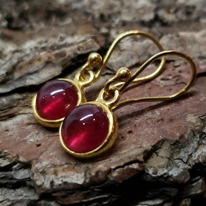 May include: A pair of gold earrings with small, round, red gemstones. The earrings have a simple, elegant design.
