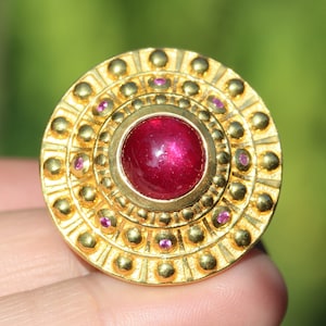 May include: A gold ring with a large, round, red ruby gemstone surrounded by smaller pink gemstones. The ring has a circular design with multiple concentric circles.