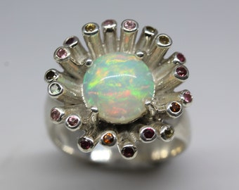 May include: A silver ring featuring a large opal gemstone encircled by a halo of smaller pink, yellow, and green gemstones.