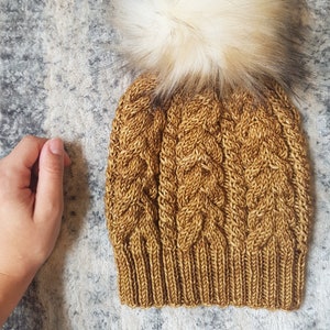 以下が含まれることがあります： 茶色のニット帽に、大きな白いフェイクファーのポンポンがついた帽子。帽子はケーブルニットの模様です。