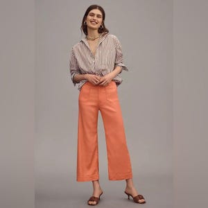 May include: A woman wearing a brown and white striped button-down shirt and orange wide-leg pants. The pants have two front pockets. The woman is also wearing brown heeled sandals and gold jewelry.