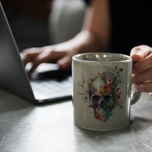 Puede incluir: Una taza de cer&aacute;mica gris con un dise&ntilde;o de calavera acuarela rodeada de flores coloridas. La taza est&aacute; siendo sostenida por una mano que lleva anillos.